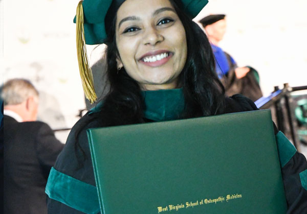 Woman smiles wearing graduation regalia holding leather bound folder.
